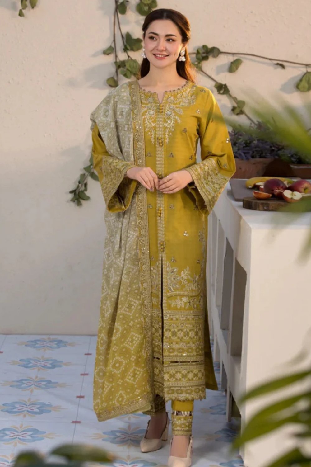 Woman in a yellow traditional outfit standing in a decorated room with plants and a table. revishstore.com revish store